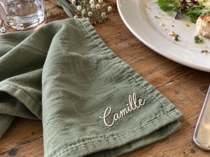 Détail généré du prénom Camille brodé près du coin d’une serviette vert sauge disposée en biais